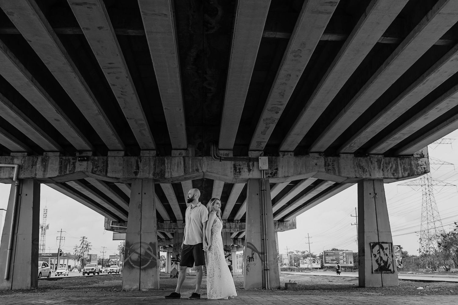 A black and white photograph showing figures under a geometric highway overpass with strong parallel lines and shadows.