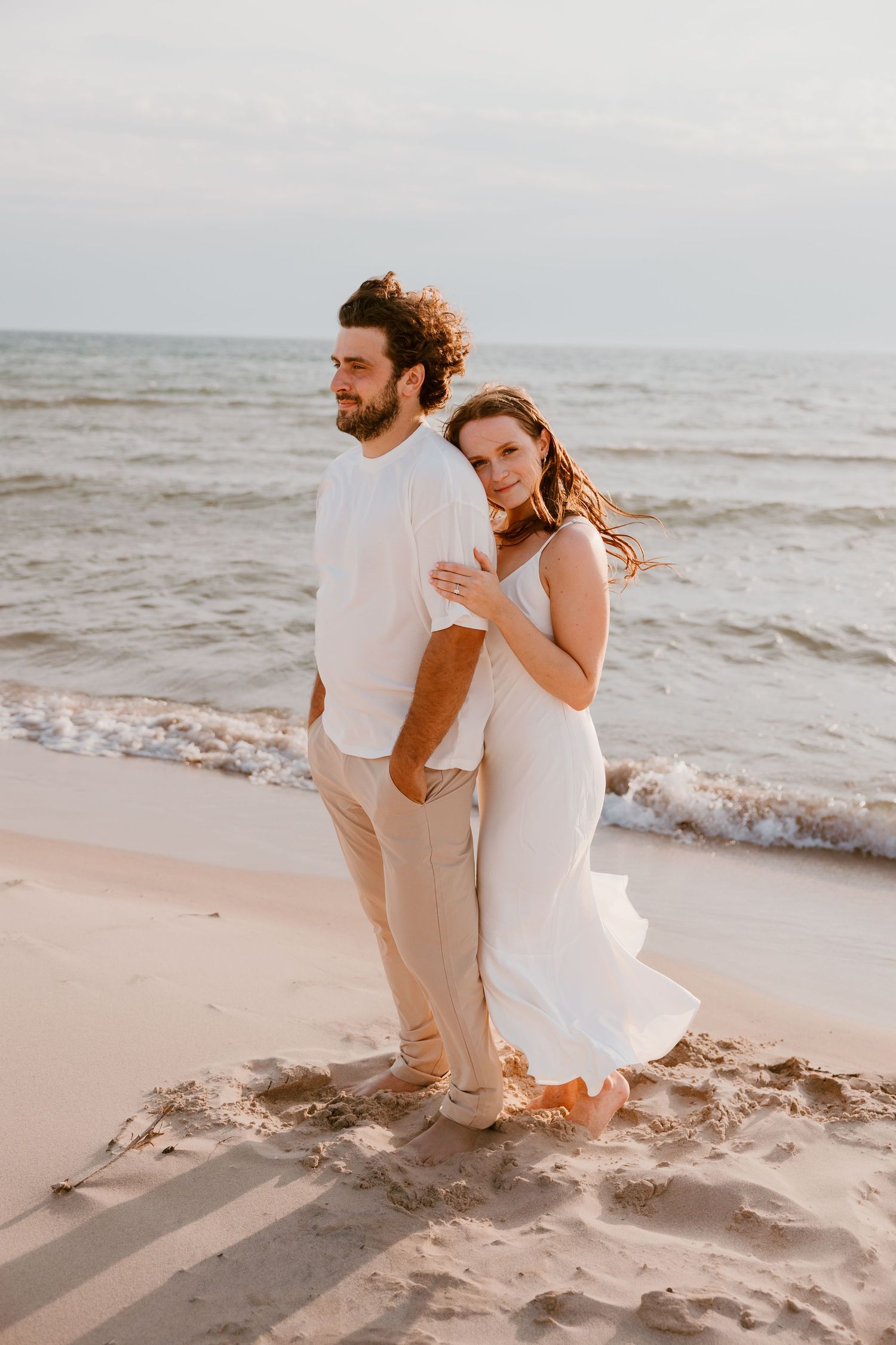 A romantic couple in white outfits stands embracing on a sandy beach at sunset with ocean waves in the background. Michigan Engagement Photo