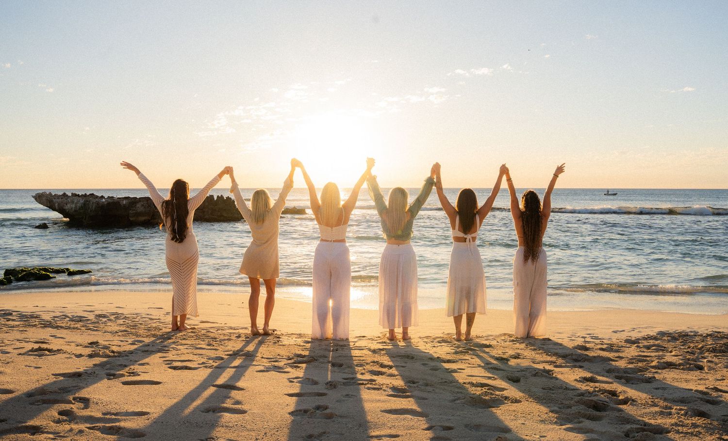Ethereal white-dressed figures dance in motion on a sunny beach at sunset with ocean waves and rocks in background.