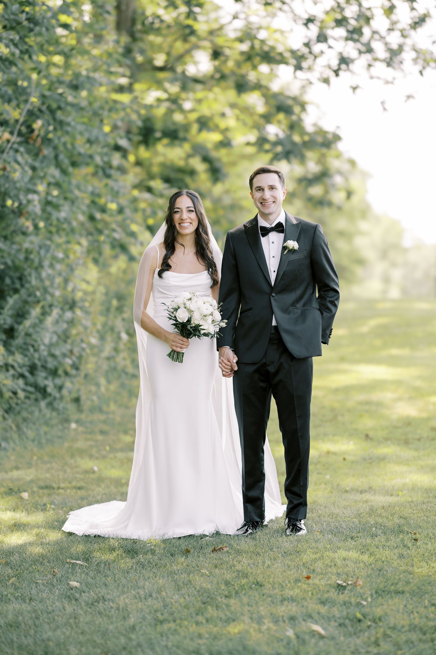 A bride and groom pose together outdoors in a sunlit garden setting for their wedding portraits.