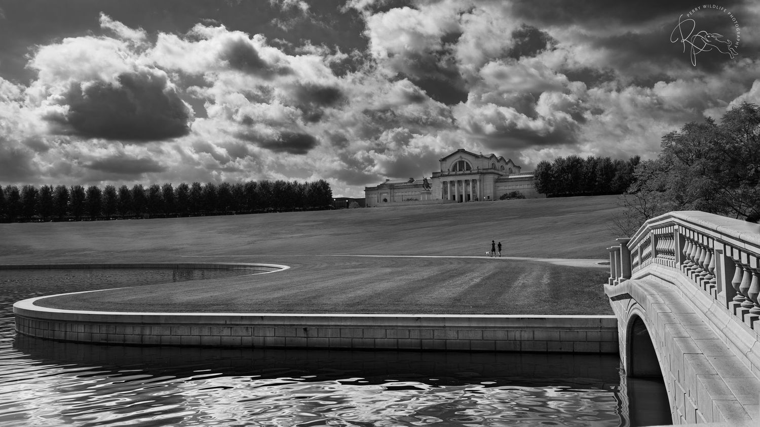 Dramatic black and white landscape of a golf course with stone bridge and historic mansion under cloudy skies.