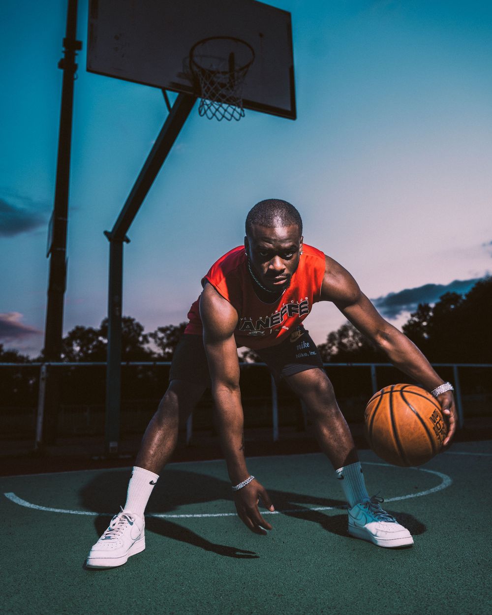 Joueur de basket-ball en maillot rouge dribblant sur un terrain extérieur au crépuscule. Florent Vause