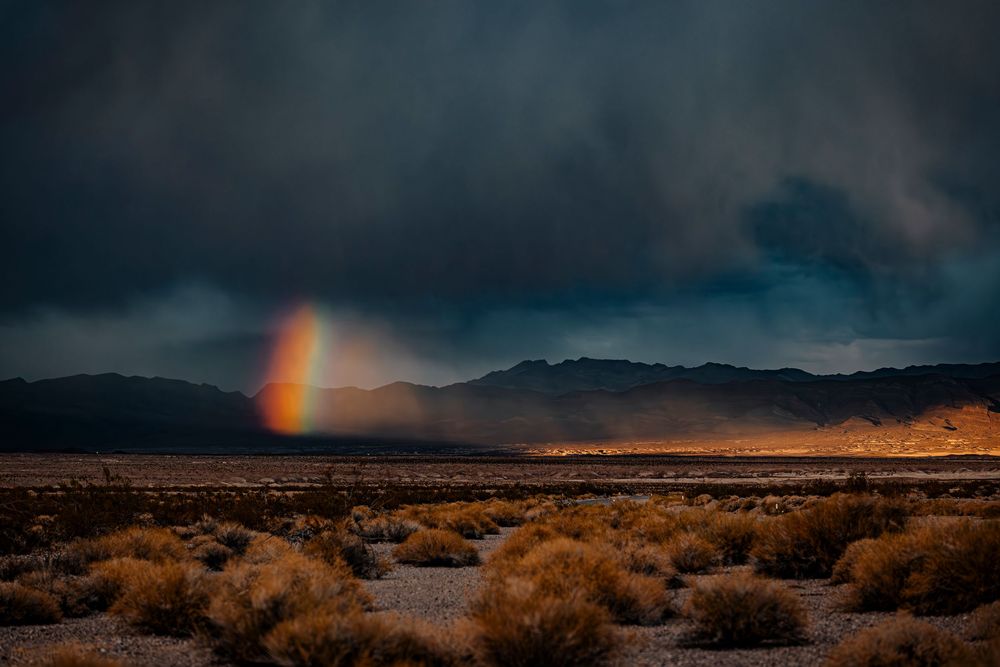 Rainbow emerging from dark storm clouds over arid desert landscape.