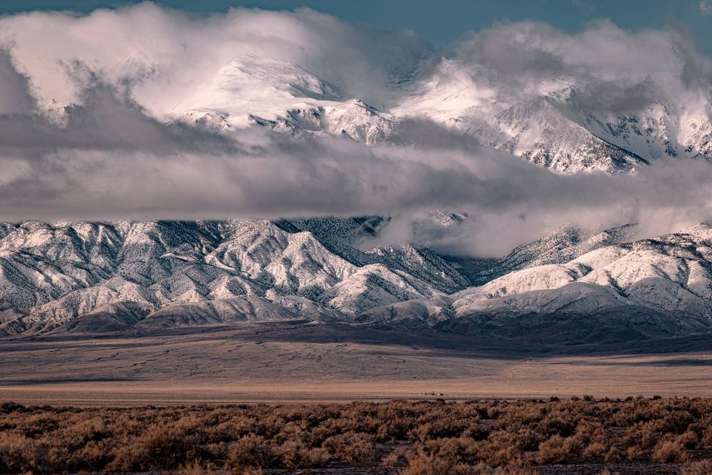 Snow-capped mountain range with dramatic clouds over arid desert flatlands in warm tones.