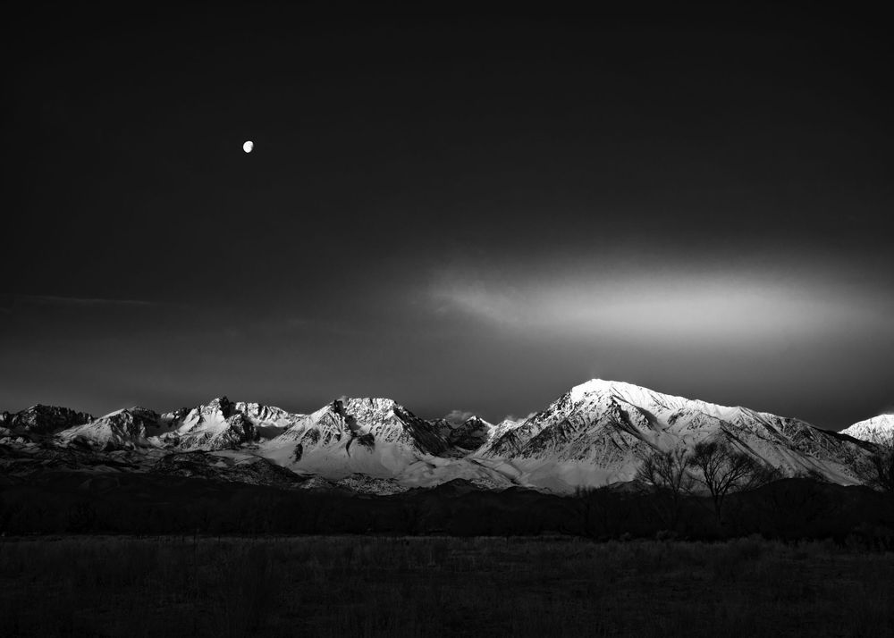 Black and white moonrise over snow-capped mountain range at dusk.