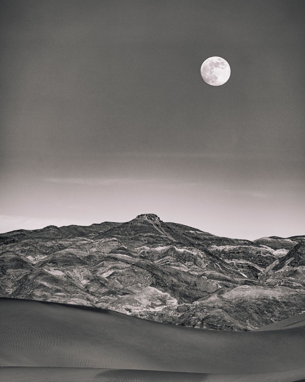 Full moon rising over rugged mountain range above sand dunes in black and white desert scene.