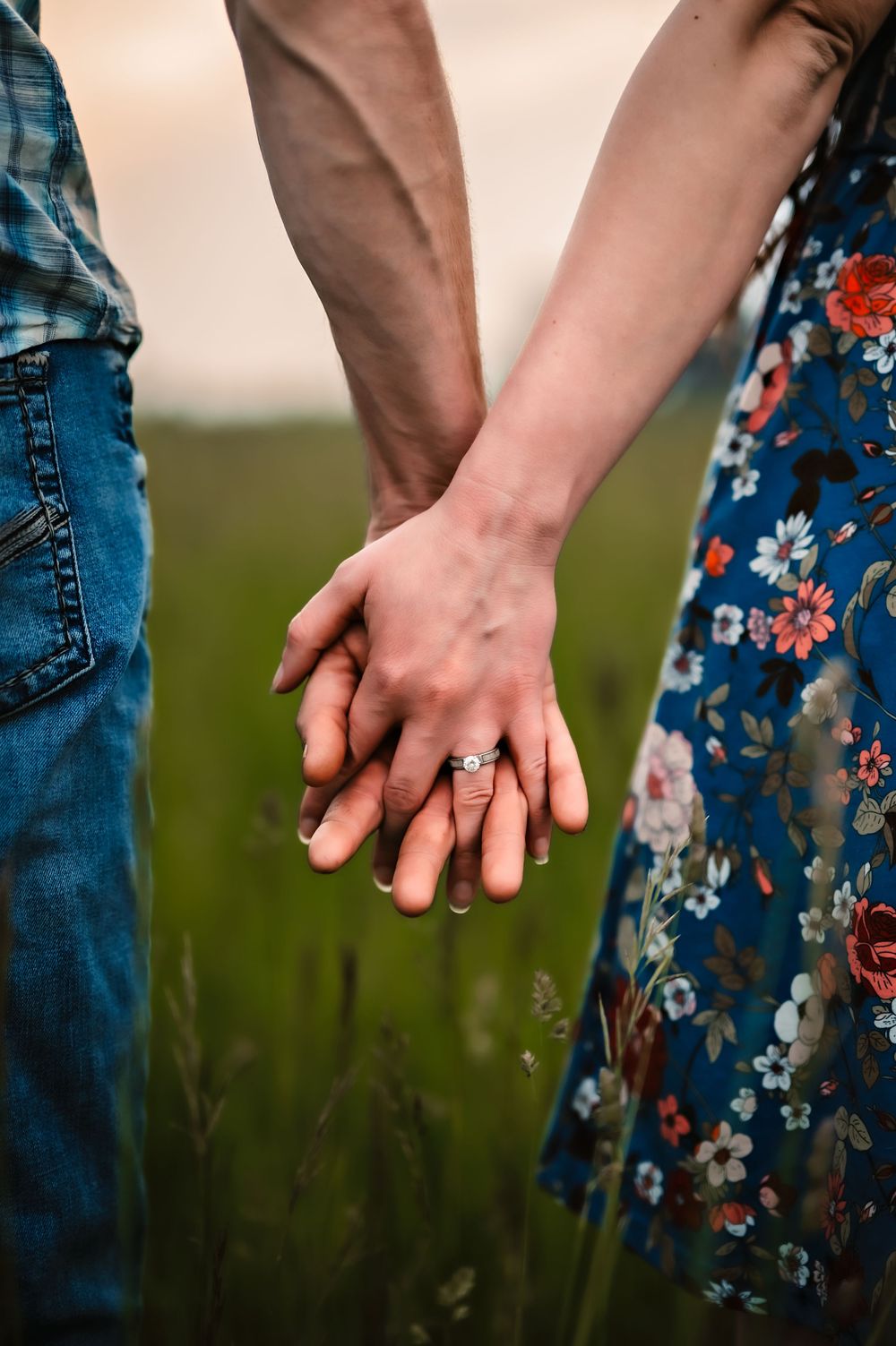 A couple holds hands in a field, wearing jeans and a floral dress, with an engagement ring visible.