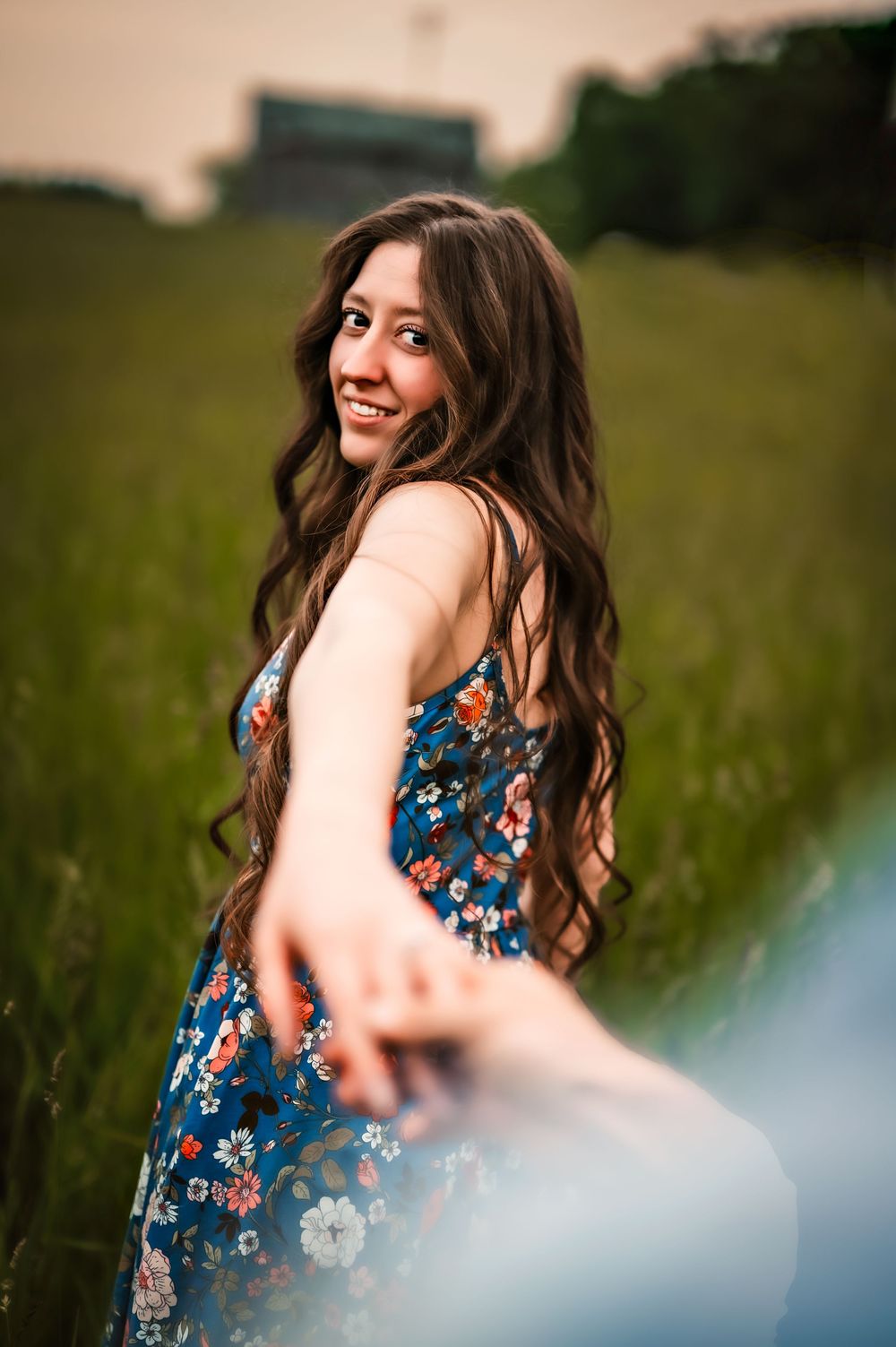 Someone wearing a blue floral dress smiles while turning back in a grassy field at golden hour.