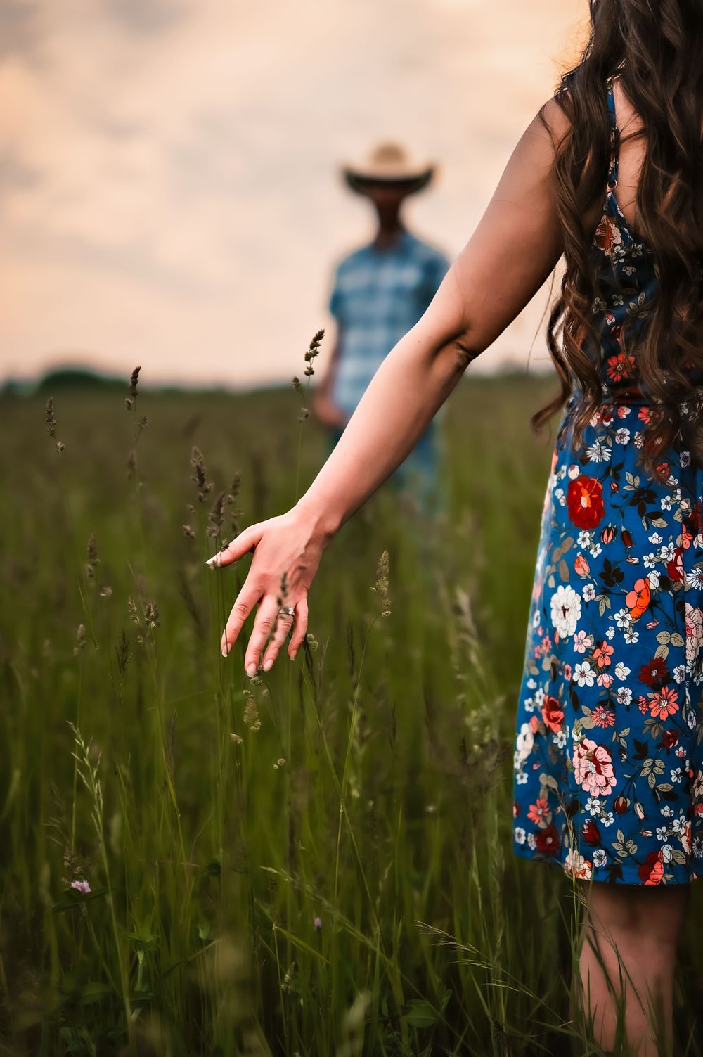 Woman in floral dress runs hand through tall grass in field at sunset with figure in cowboy hat in background.