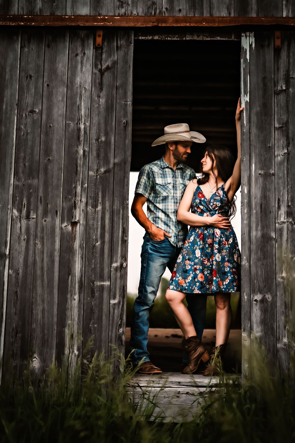 A romantic country couple in western attire poses together in a rustic barn doorway.