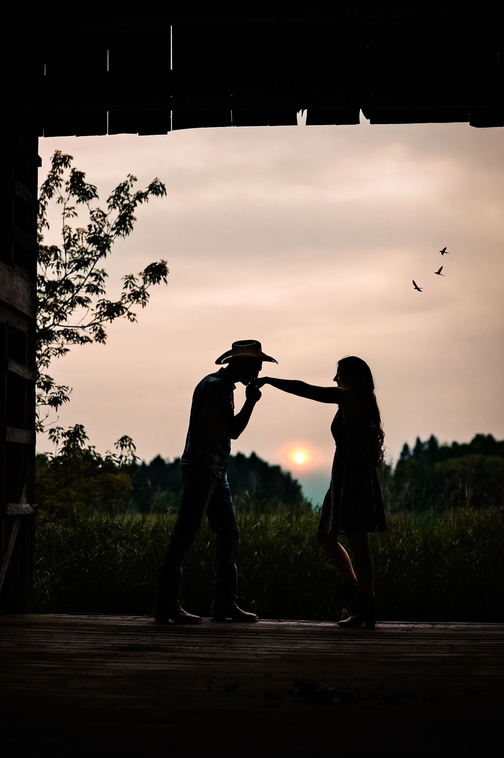 A romantic silhouette of two people dancing against a sunset sky with birds flying in the background.