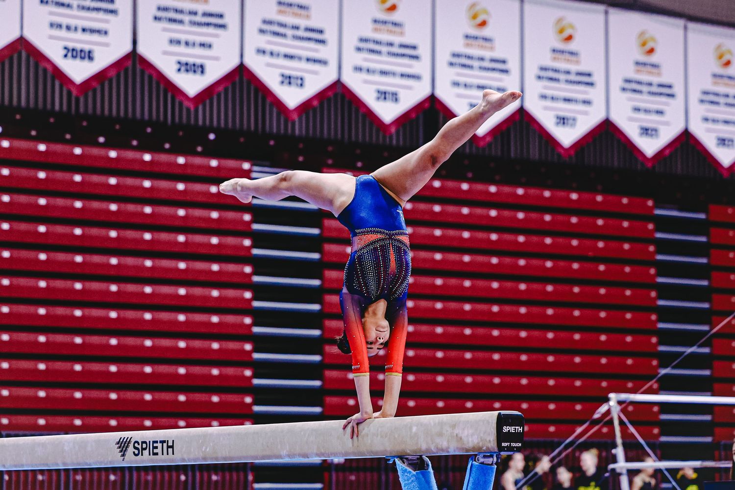 A gymnast performs a split leap in mid-air during a competition, wearing a navy blue leotard.