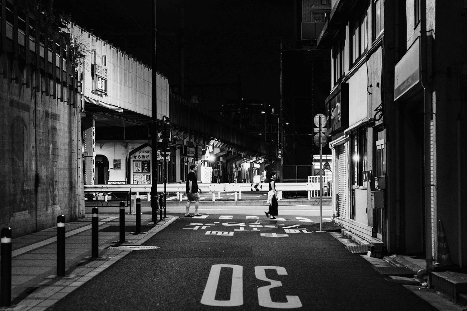Empty urban street at night with road markings and illuminated buildings in black and white.
