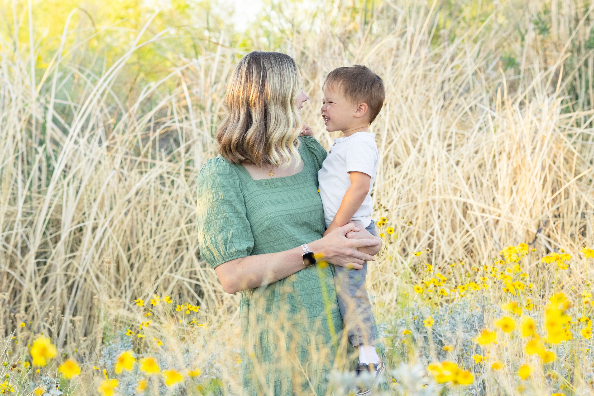 Mom holding toddler son, smiling at each other in bushes of yellow flowers in Gilbert, AZ at the Riparian Preserve.