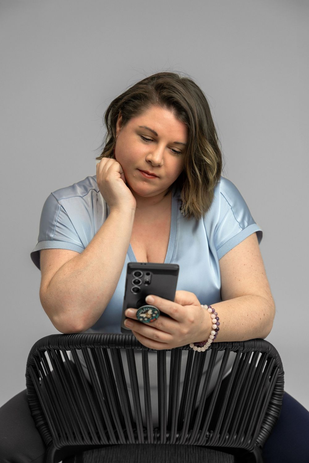 A person wearing a light blue shirt leans on a black chair while looking at a phone against a gray backdrop.