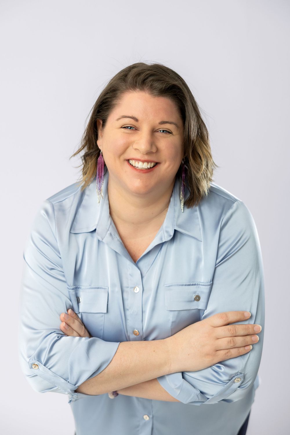 A woman wearing a light blue button-down shirt smiles and poses against a light gray background in studio portraits.
