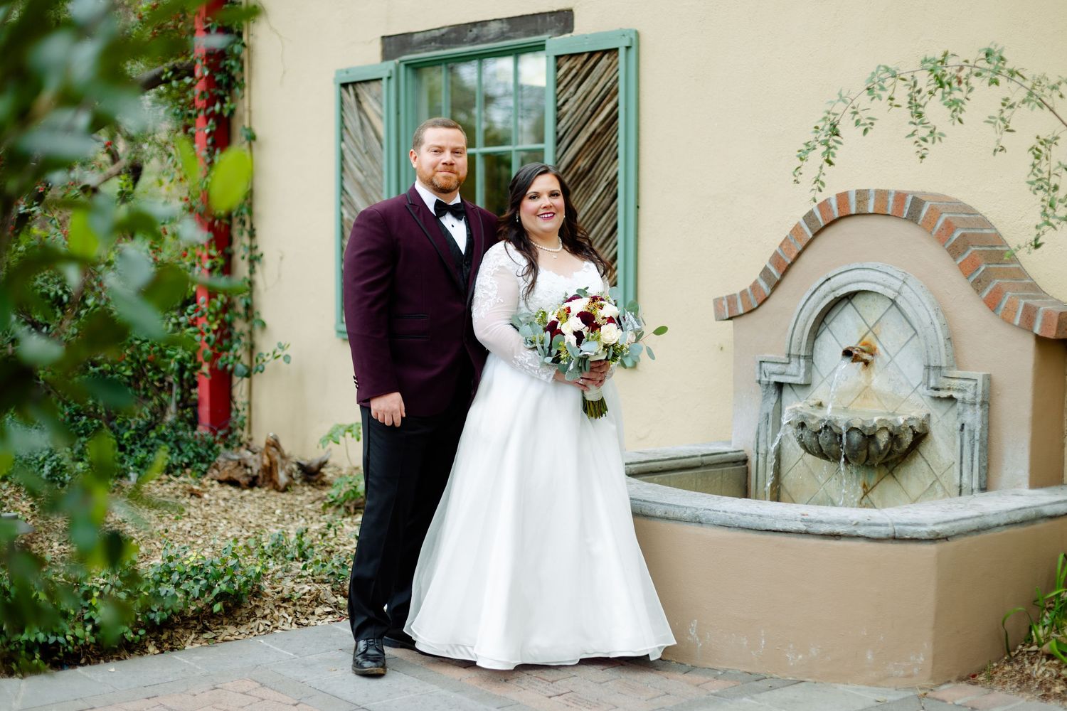 A wedding couple poses in front of a stucco wall with a decorative water fountain and green shuttered window.