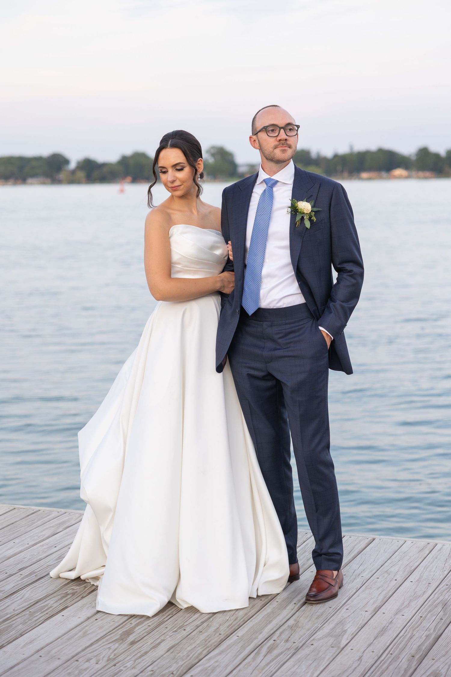 A couple in wedding attire stands on a wooden dock overlooking a serene waterfront at sunset by Rachel Martinez Photography in Metro Detroit