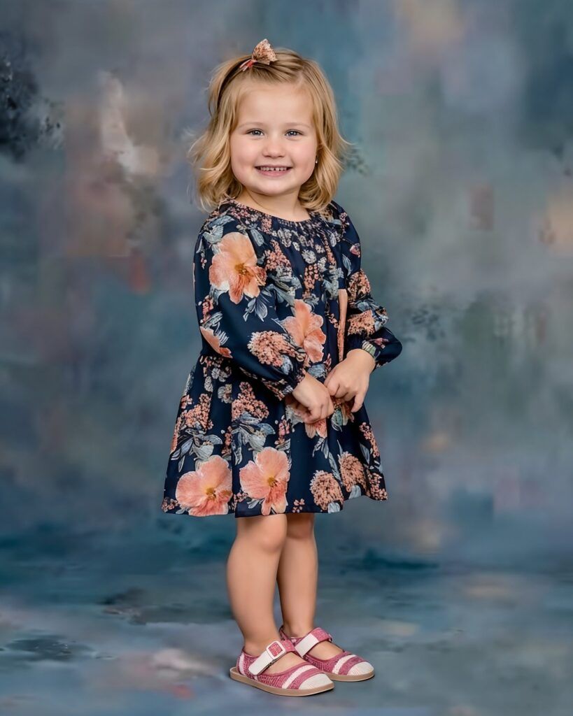 A child wearing a navy floral dress with peach roses and pink sandals poses against a mottled blue-gray backdrop.