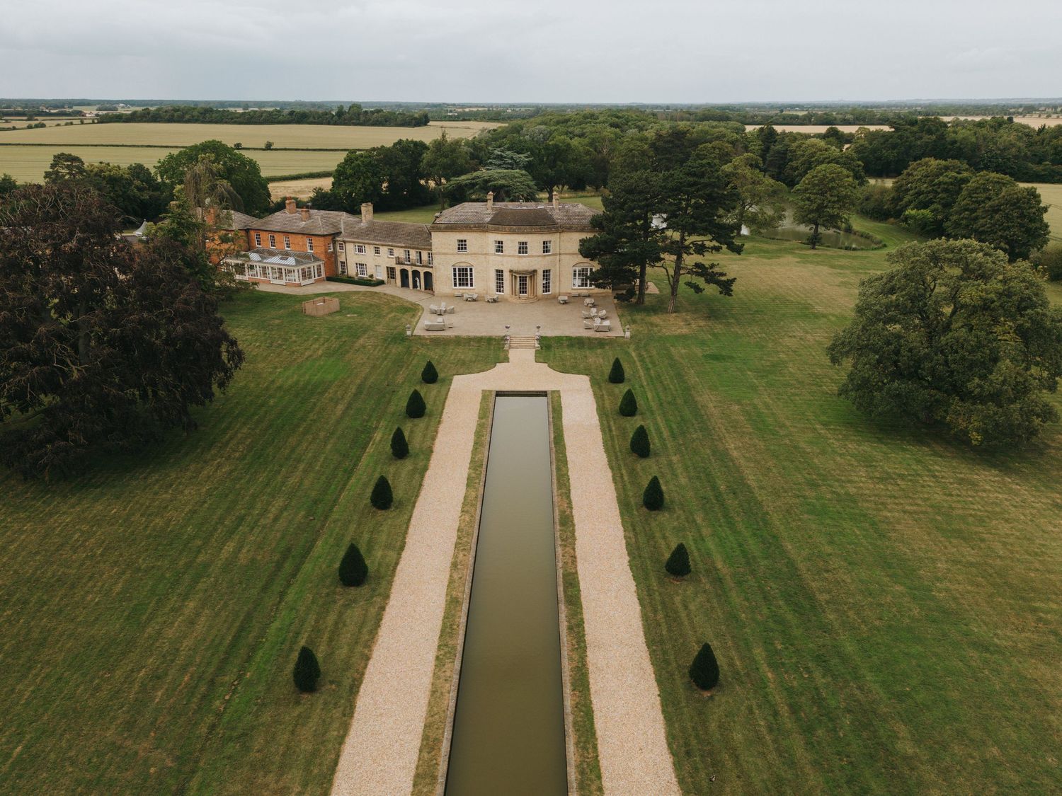 Aerial view of a grand country estate with manicured lawns and a long rectangular reflecting pool leading to the mansion.