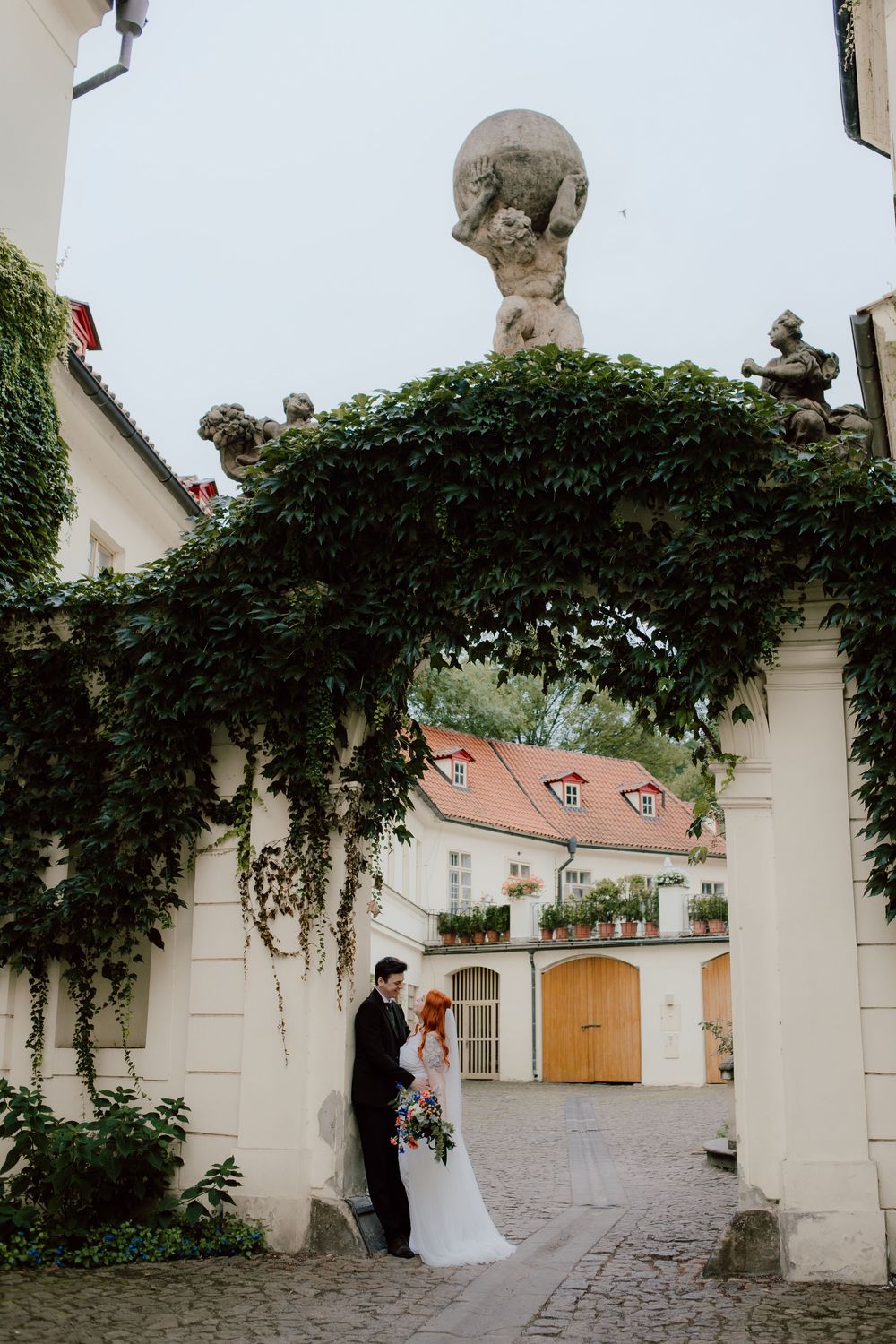 A romantic wedding couple stands under an ivy-covered archway in a charming European courtyard setting.