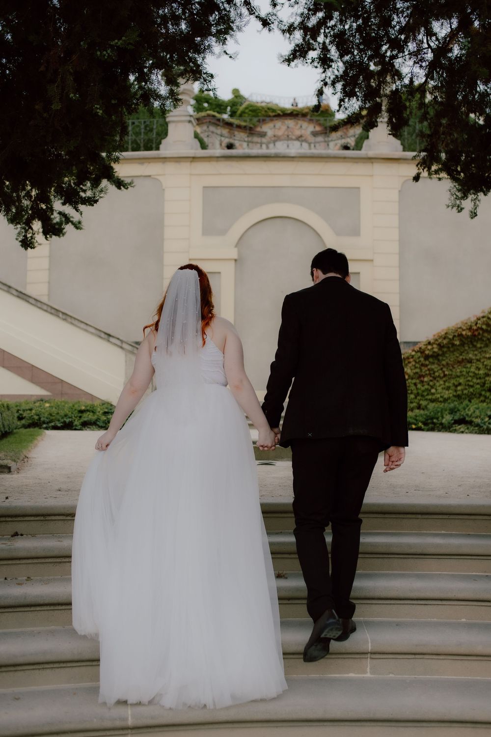 A bride and groom ascend white stone steps while holding hands against a classical architectural backdrop.