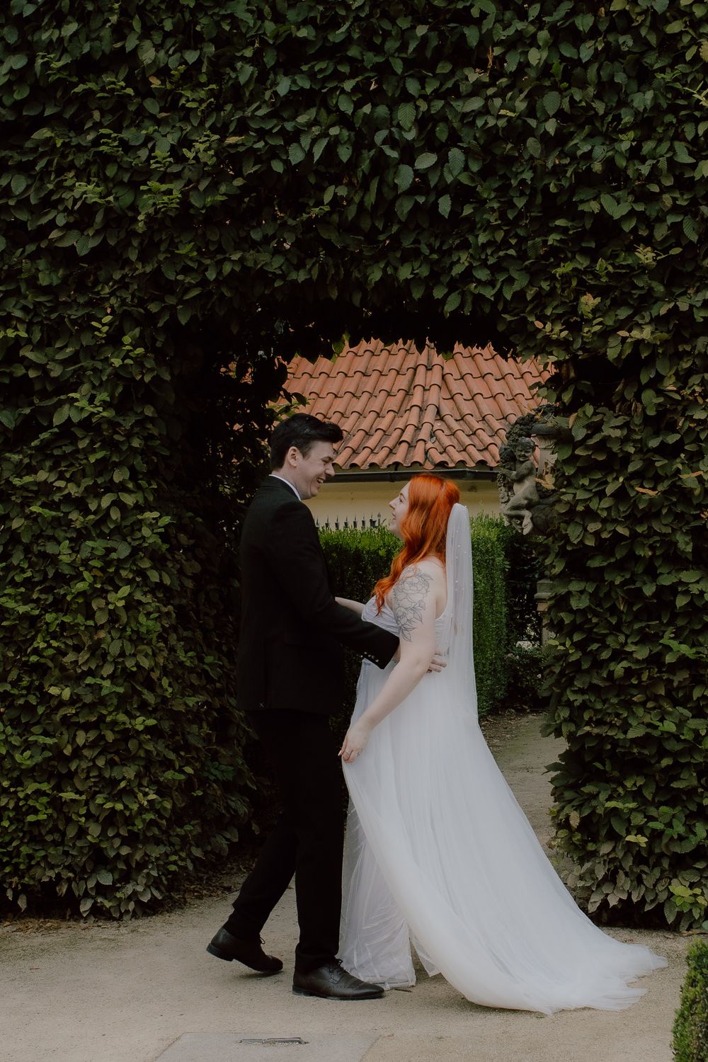 Wedding couple holding hands in romantic garden walkway framed by tall green hedges and terracotta roof in background.