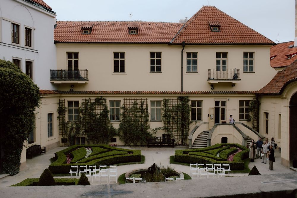 Elegant courtyard garden featuring white chairs and manicured hedges surrounded by historic European architecture with terra cotta roof.