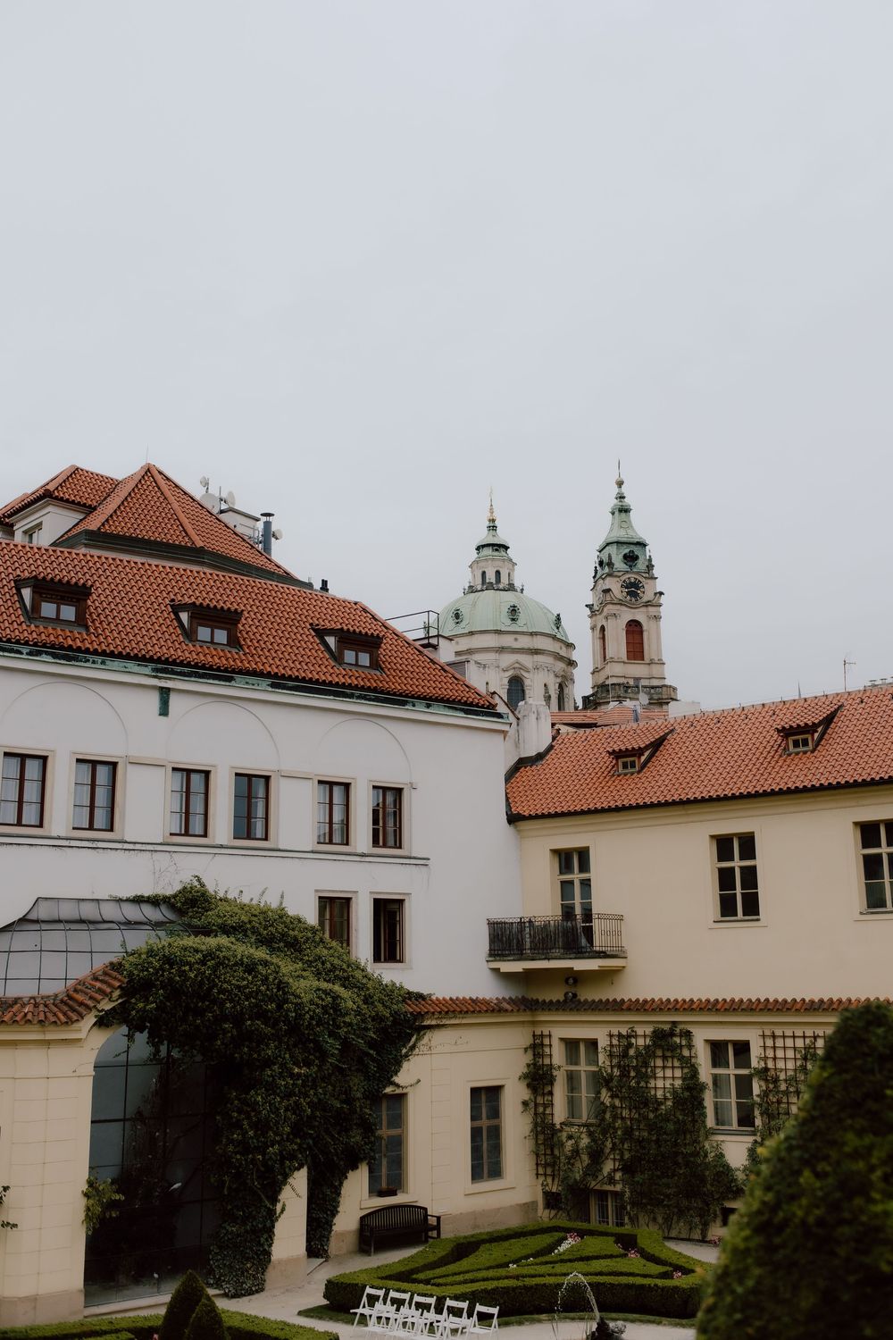 Panoramic view of historic Prague building complex with baroque church towers and red tiled roofs in cloudy weather.