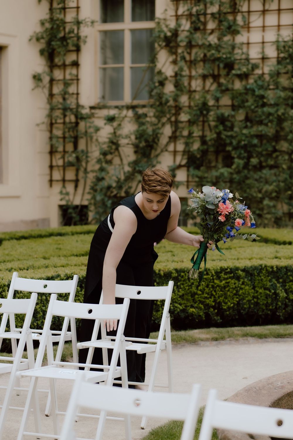 Wedding staff placing decorative floral bouquet on white folding chairs at an outdoor garden ceremony setup.