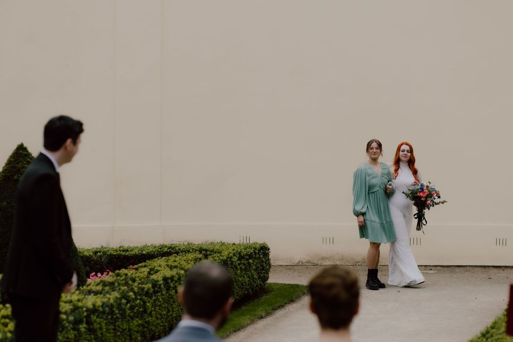 Outdoors wedding scene with people gathered along a manicured hedge and white wall.