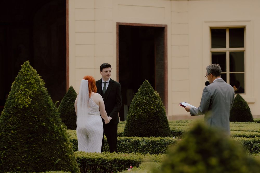 Two people stand in formal attire outside a building with manicured bushes at an elegant venue.