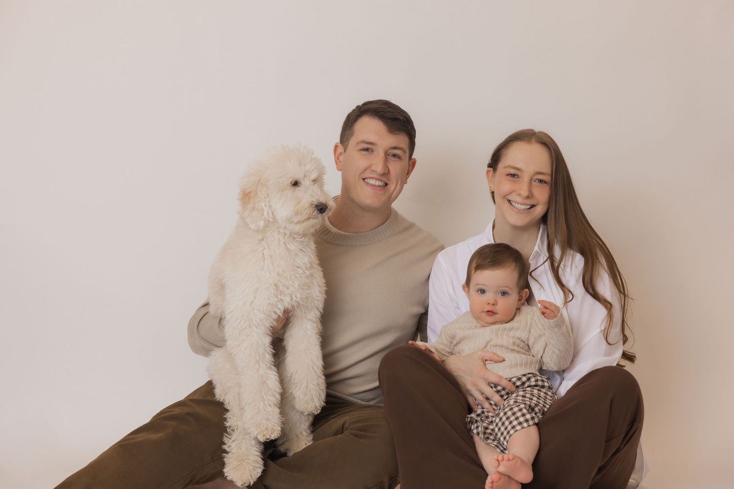 A family poses for a series of casual portraits with their white dog on a neutral background.