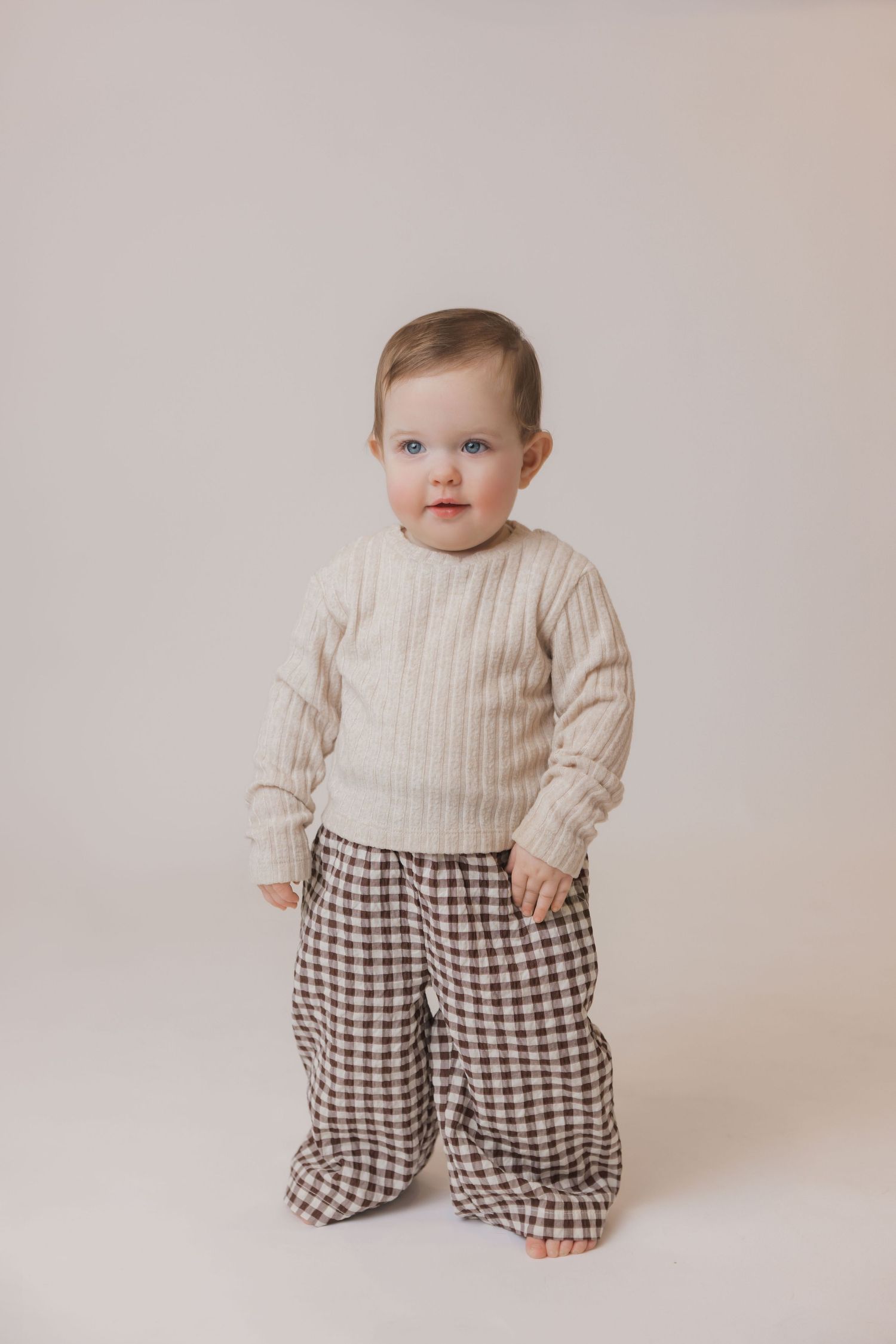 A baby wearing a cream sweater and checkered pants stands against a neutral backdrop in a series of progressive poses.