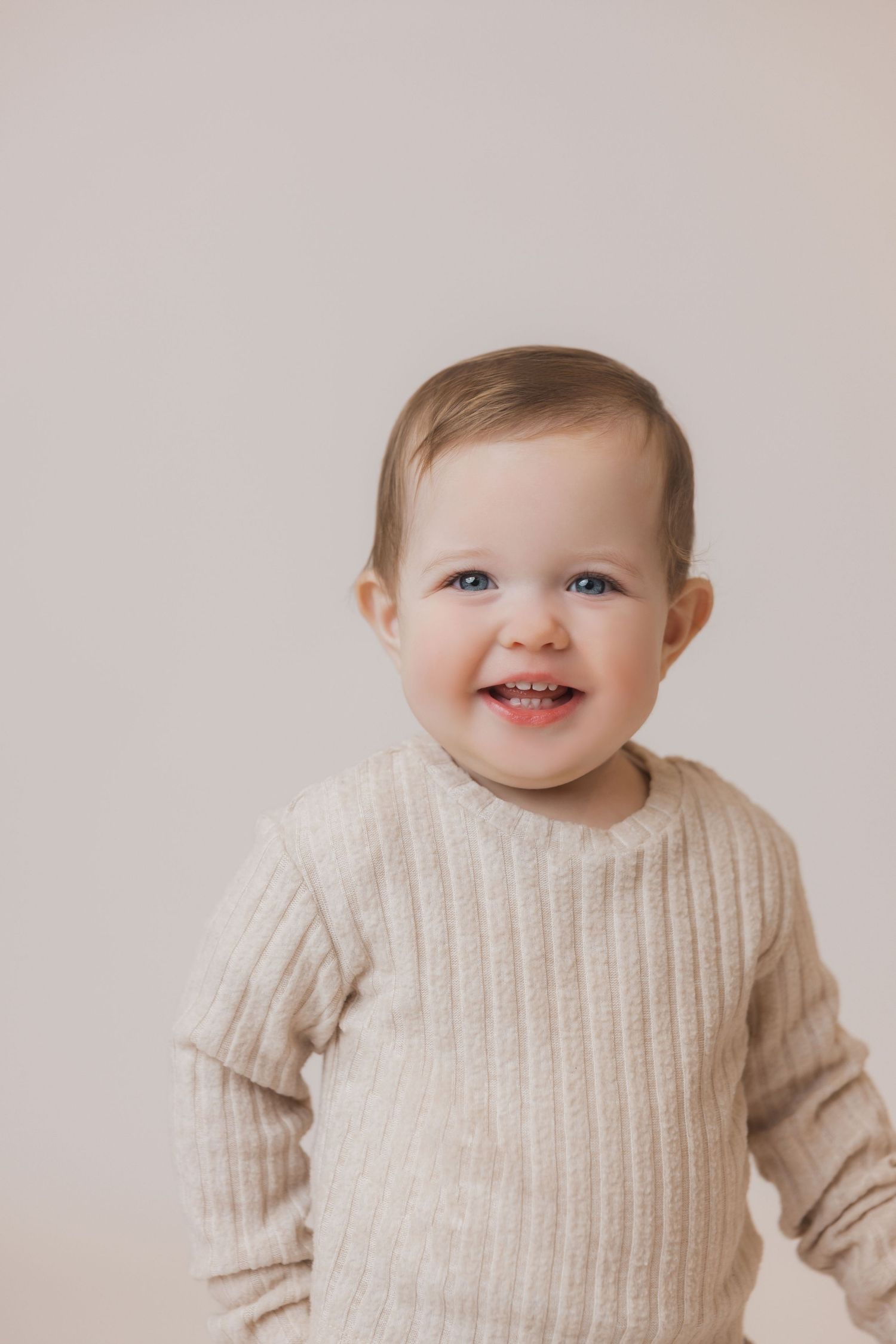A happy baby wearing a cream colored ribbed sweater smiles against a neutral background.