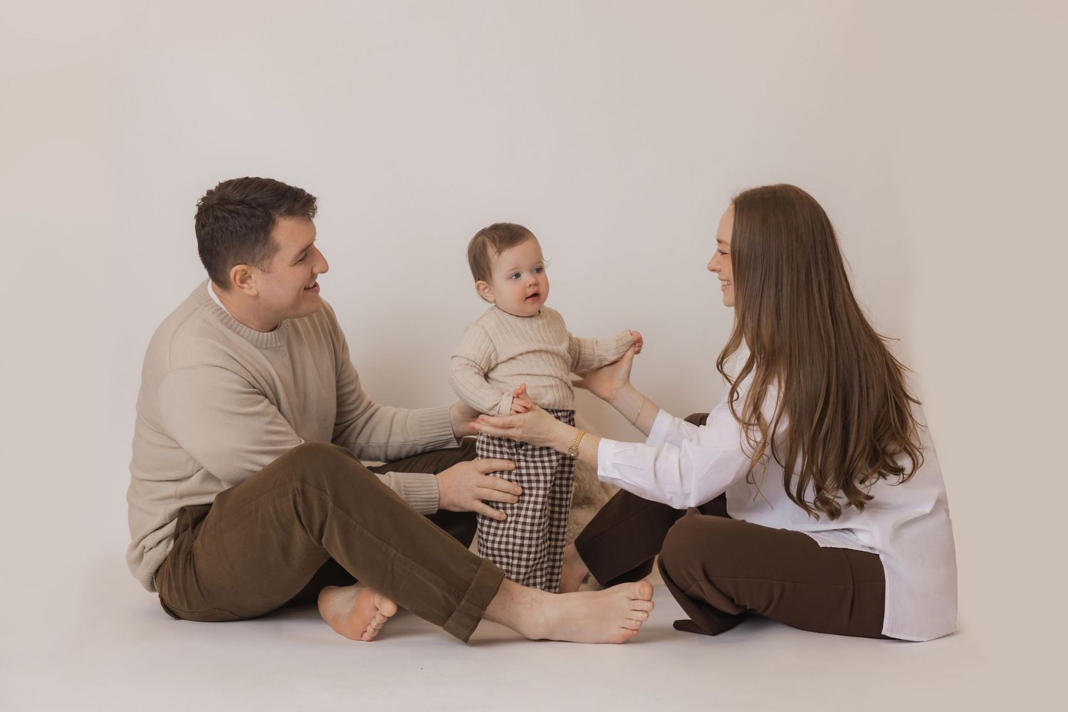Family wearing neutral colors lies together on beige backdrop in studio portrait session.