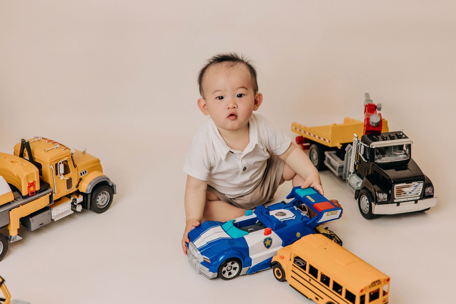 A child in a white shirt sits on the floor surrounded by colorful toy vehicles including trucks, buses and police cars.