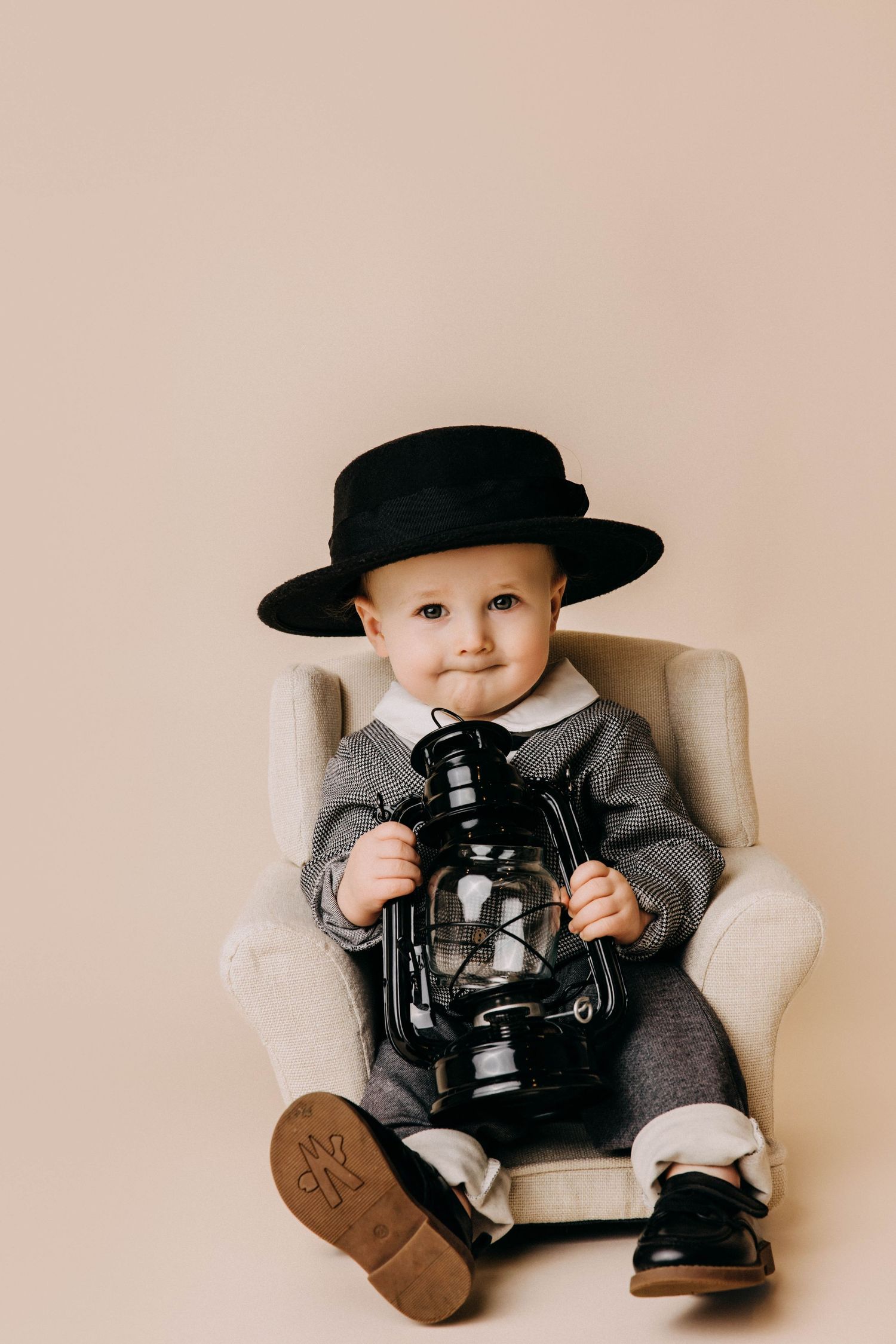 Baby in formal attire holds vintage lantern while seated in cream armchair.