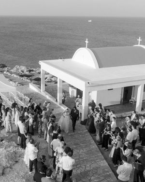 A crowd gathers outside a white Mediterranean-style church with crosses on its domed roof overlooking the sea.