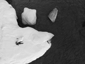 An aerial view shows two people skiing down a steep snowy slope with dramatic cliffs and dark water below.