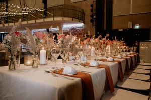 Elegant wedding reception table setup with white linens, terracotta runners, candles and floral centerpieces in indoor venue.
