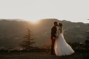 A couple in wedding attire silhouetted against a golden sunset on a mountain overlook with evergreen trees in view.