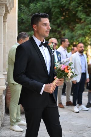 Wedding party members in formal wear with bouquets pose in an outdoor courtyard setting.