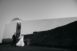 A black and white photo of a couple in wedding attire walking near a modern architectural structure.