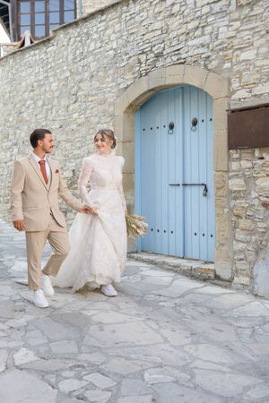 Two people in formal attire walking along a stone wall with light blue doors in Mediterranean setting.