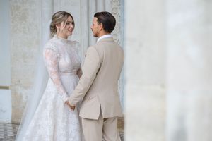 Bride and groom share tender moments in front of light stone wall wearing elegant lace gown and tan suit.
