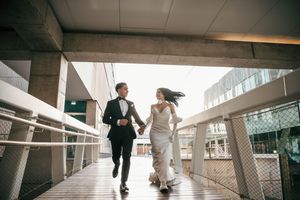 A couple in wedding attire runs joyfully together on a modern covered walkway with blowing wind.