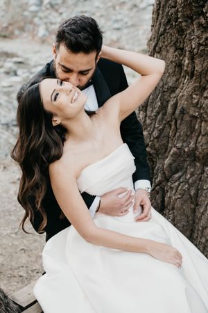 A couple in wedding attire share an intimate moment while leaning against a tree trunk outdoors.