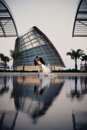 A romantic wedding couple poses against a modern architectural backdrop with palm trees and reflective surfaces.