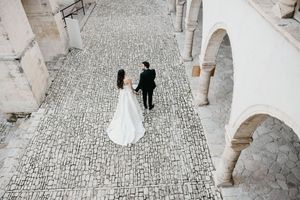A couple in wedding attire poses on a cobblestone street surrounded by elegant stone archways.